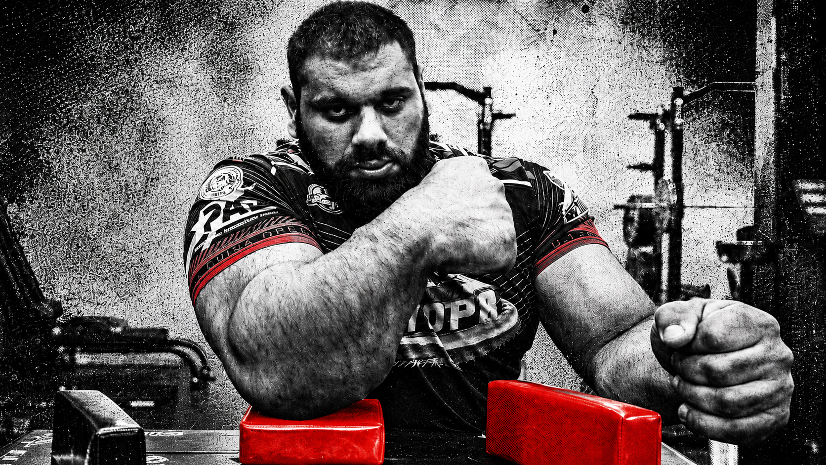 Levan Saginashvili poses at an arm wrestling table in a high-contrast black-and-white gym image with red accents.